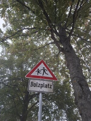 Bild mit ausf&uuml;hrlicher Beschreibung vergr&ouml;&szlig;ern: Das Bild zeigt ein dreieckiges Verkehrszeichen mit spielenden Kindern und rotem Rand. Darunter ist der Schriftzug Bolzplatz zu lesen.